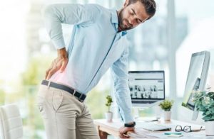 An adult male wearing a light blue dress shirt and light brown pants with a black belt and watch leans forward on his work desk with one hand as he places his other hand on his lower back and looks back in pain.