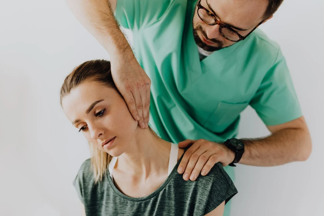 A close up of a woman wearing a dark colored tank top laying on a chiropractic table while the hands of a chiropractor wearing a wedding ring press down on her back near her shoulder.