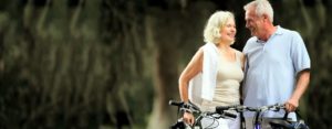 An image of an older couple standing next to bicycles and smiling. The woman is blonde and wears a tan tank top with white jeans and a sweater draped over her shoulders while the man wears a light blue polo and tan pants while he looks lovingly at the woman.