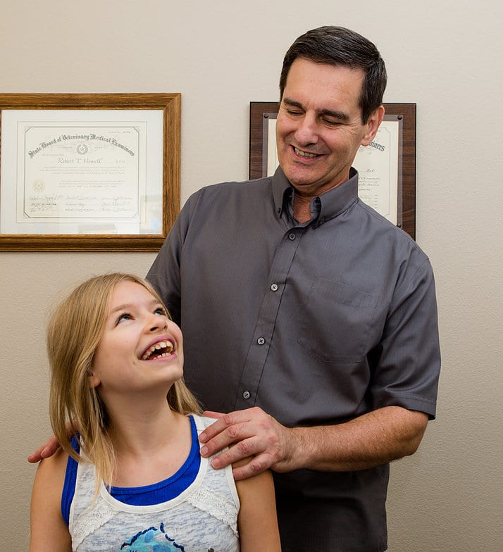 Dr. Robert T. Howell, DVM, DC, FACO standing in front of a wall featuring his certificates while wearing a gray button up shirt and black slacks. He has dark hair and is smiling, standing with his hand on the shoulder of a happy young girl.