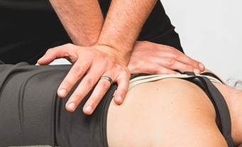A close up of a woman wearing a dark colored tank top laying on a chiropractic table while the hands of a chiropractor wearing a wedding ring press down on her back near her shoulder.